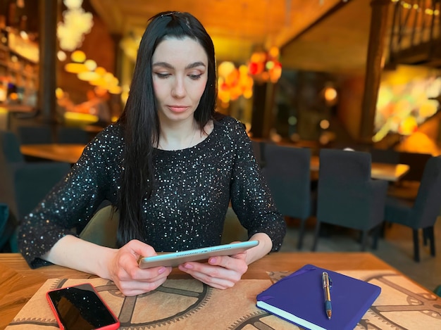 A girl with dark hair is working in a cafe with a tablet, a phone and a notebook are nearby. Coworking, workflow organization. Business concept.