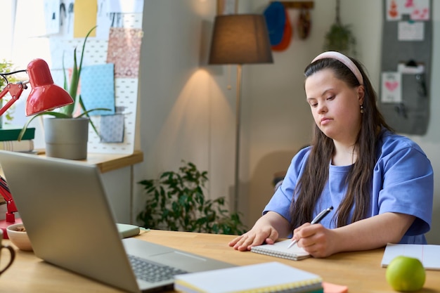 Girl with down syndrome making notes in notepad during online lesson