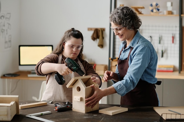 Photo girl with down syndrome using drill to make birdhouse together with master in workshop