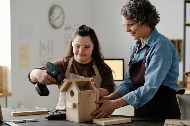 Photo girl with down syndrome using drill to make birdhouse with woman helping her with it in workshop