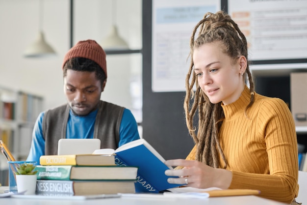 Girl with dreadlocks reading book while sitting in the library with her classmate they preparing for exam