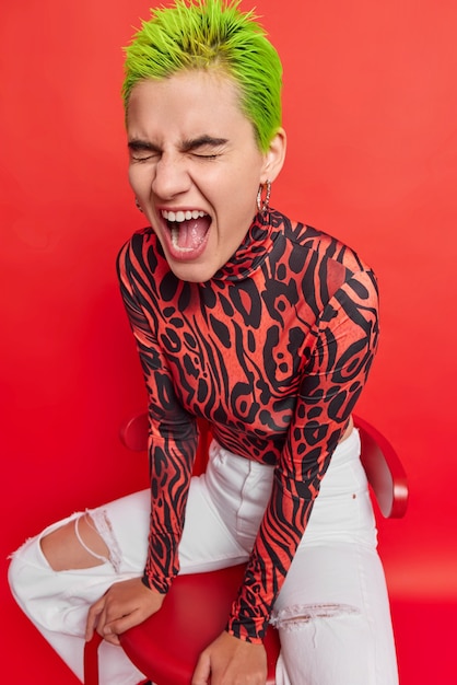 Photo girl with dyed hair poses on chair exclaims loudly dressed in stylish clothes on red