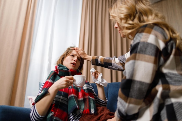 Girl with the flu drinks hot tea while her mother checks her forehead for a fever