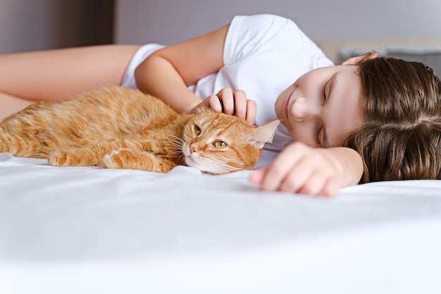 A girl with a ginger cat lies on a white bed. A young girl is stroking a ginger cat while lying on a white sheet