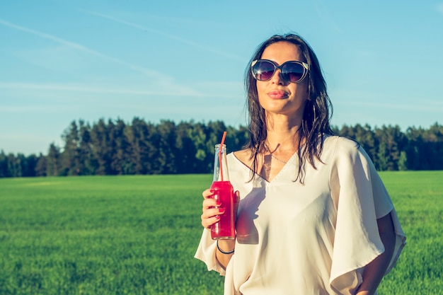 девушка в очках и вьющиеся волосы улыбаться и пить алкогольный или безалкогольный красный коктейль через соломинку из бутылки в солнечный летний день, улыбаясь на открытом воздухе