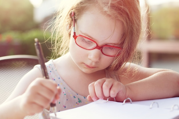 眼鏡をかけた女の子がノートに書き込みます。日当たりの良い柔らかい背景。屋外で宿題をする