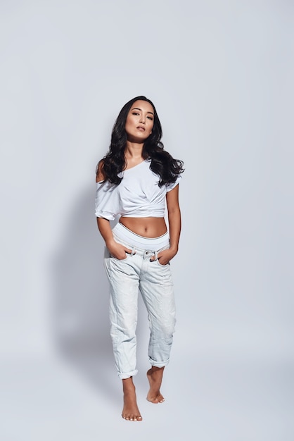 Girl with a great style. Full length of beautiful young woman keeping hands in pockets and looking at camera while standing against grey background