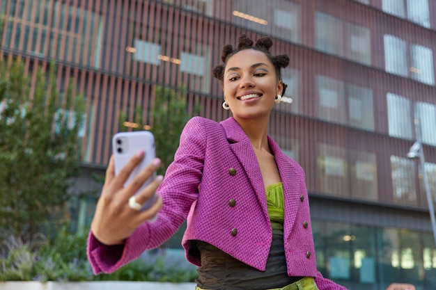 girl with hair buns looks positively at smartphone camera speaks on video call smiles happily makes selfie against modern city building connected to wireless internet