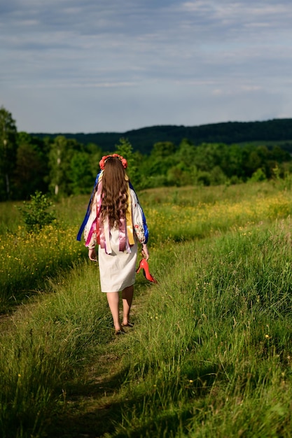 Девушка, повернувшаяся спиной и одетая в украинскую национальную одежду, ходит босиком по полю в венке с лентами на голове.