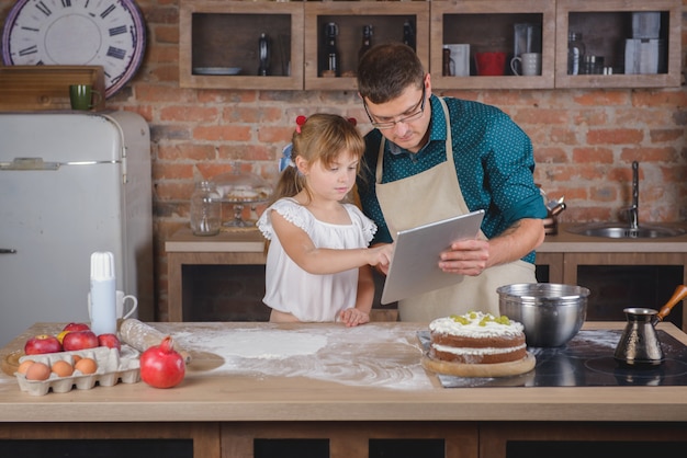 A girl with her father in the kitchen with a digital tablet are preparing dinner