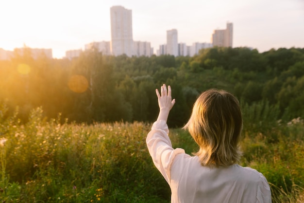 Foto una ragazza con la mano alzata si trova su una montagna nella fotocamera guardando la città in lontananza al tramonto