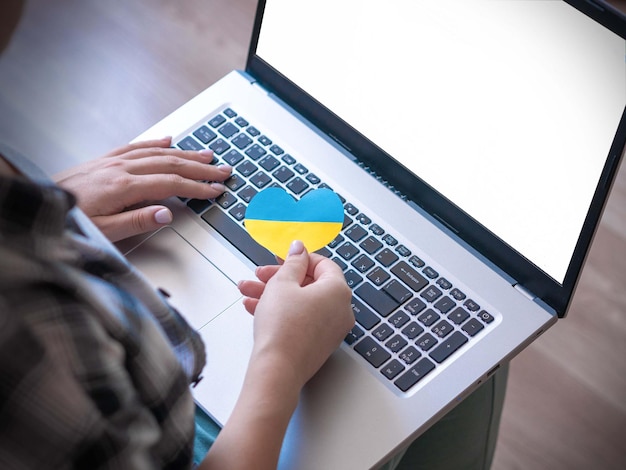 A girl with a laptop holding a yellow and blue heart of the flag of Ukraine in front of the keyboard