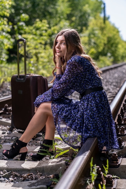 A girl with a large travel suitcase sat down to rest on the rails that are laid through a dense green forest. Travel alone