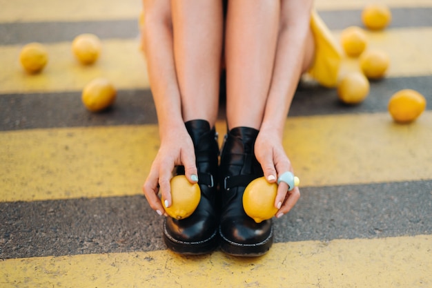 Девушка с лимонами в желтой рубашке, шортах и черных туфлях сидит на желтом пешеходном переходе в городе