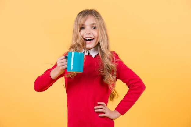 Girl with long blond hair in red sweater hold mug. Child smile with blue cup on orange background. Health and healthy drink. Thirst, dehydration concept. Tea or coffee break.