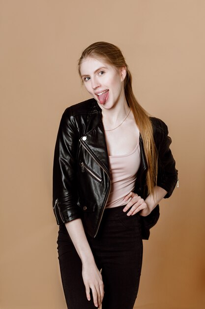 A girl with long blonde hair shows her tongue to the camera on a beige background