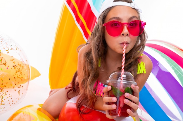 Girl with long crucified hair in sunglasses and swimsuit sits on inflatable mattresses drinks fresh fruit cocktail isolated on a white background