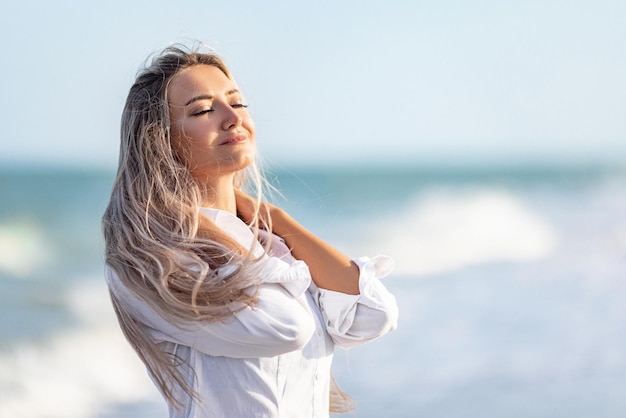 A girl with long hair in a bluish swimsuit and a shirt goes through her hair on the seashore