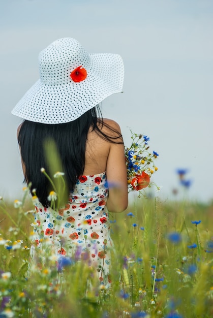 girl with long hair in a dress and a white hat with poppy flower stands with in a blossoming meadow with a bouquet of wildflowers.