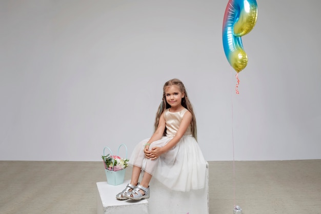 Ragazza con i capelli lunghi in abito festivo seduto vicino a bouquet di fiori sfondo bianco studio