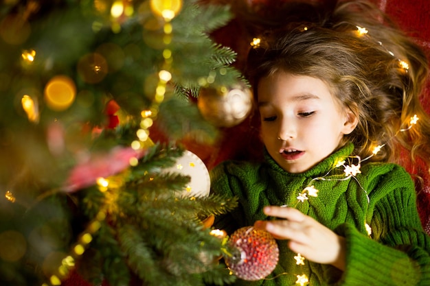 긴 머리와 화환을 가진 소녀는 따뜻한 니트 스웨터에 장난감이 있는 크리스마스 트리 아래 빨간 격자 무늬 위에 누워 있습니다. 크리스마스, 새해, 아이들의 감정, 기쁨, 기적에 대한 기대와 선물