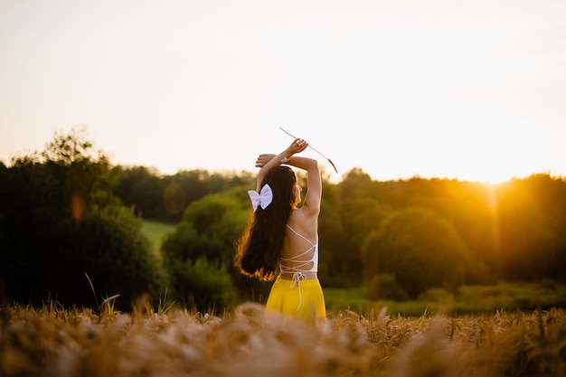 A girl with long hair stands in a yellow skirt in a field with spikelets and looks at the sun