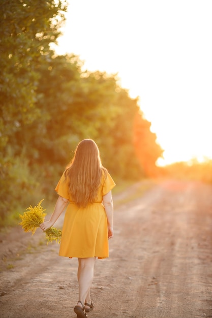 A girl with long hair walks in the field with a bouquet of yellow flowers at sunset