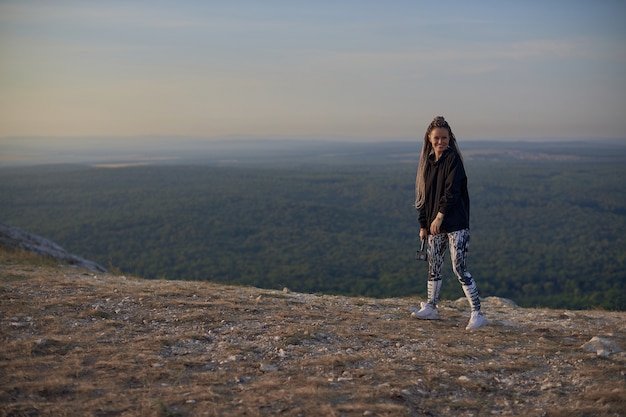 A girl with long pigtails in sportswear and white sneakers stands on top of a mountain overlooking t...
