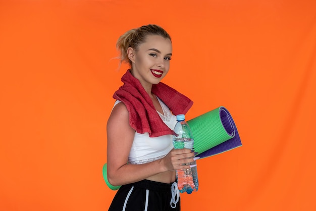 Girl with makeup with a beautiful figure holds a mat and a water bottle near the wall