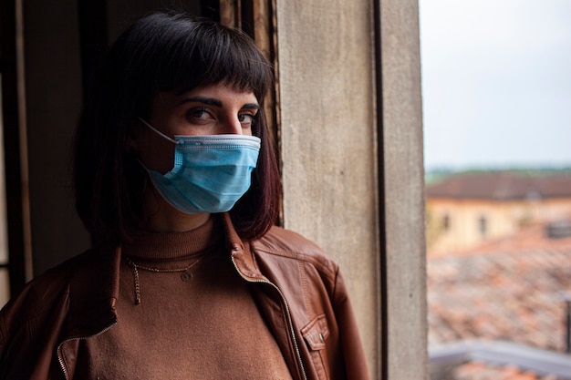 Girl with medical mask at window in her home during covid quarantine period