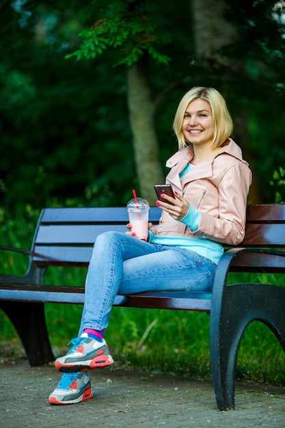 Girl with milkshake talking on phone sitting on bench at street in summer