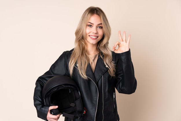 девушка с мотоциклетным шлемом на бежевой стене показывает знак ОК пальцами