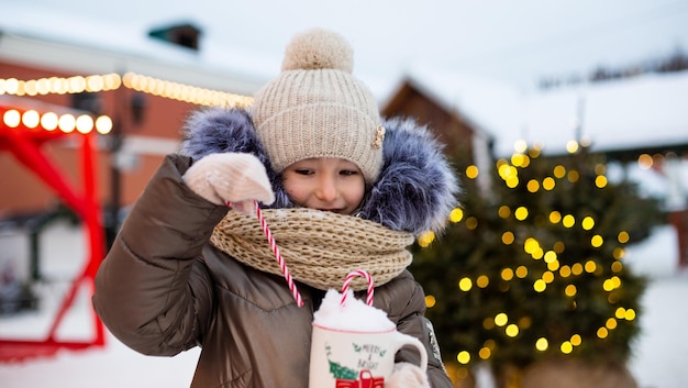 Girl with mug with snow candy cane and inscription Merry and Bright in her hands outdoor in warm clothes in winter at festive market Fairy lights garlands decorated snow town for new year Christmas