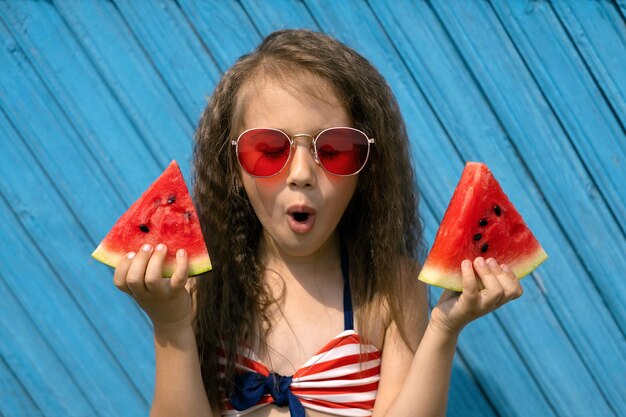 A  girl with an open mouth and a surprised face holds  pieces of watermelon in her hands