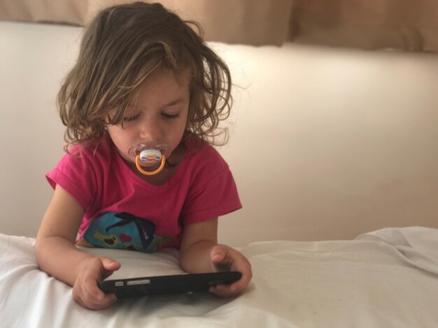 Photo girl with pacifier in mouth while using phone on bed