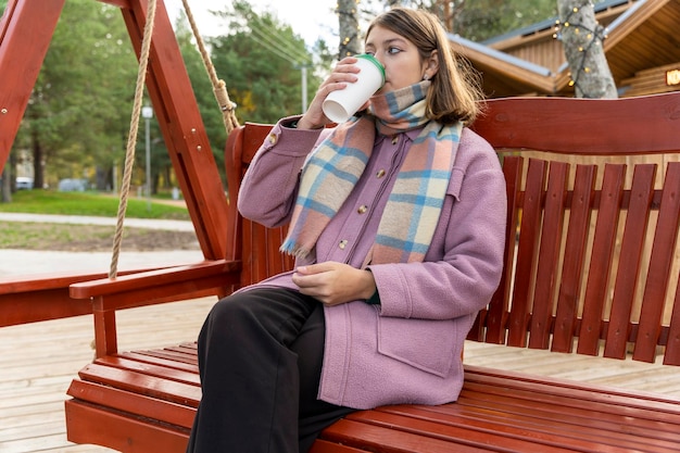 Girl with paper cup of coffee swings on bench in the park