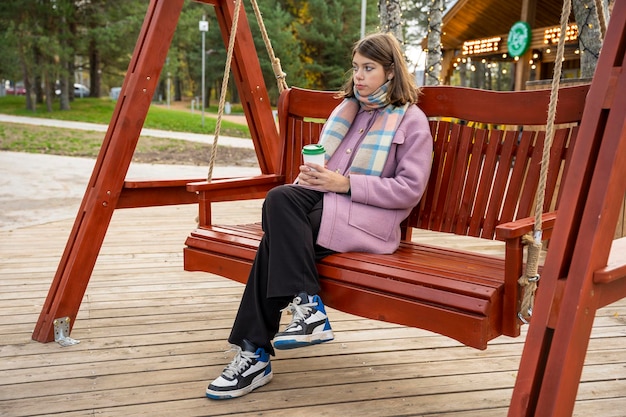 Girl with paper cup of coffee swings on bench in the park