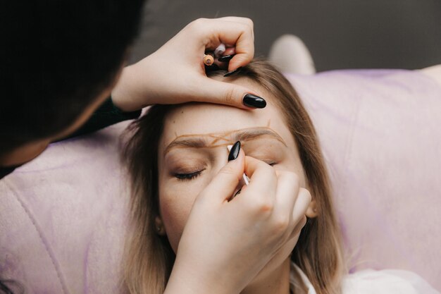 Girl with permanent makeup on her eyebrows The makeup artist makes markings with a brown pencil for eyebrow tattooing