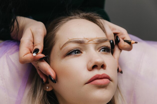 Girl with permanent makeup on her eyebrows the makeup artist
makes markings with a brown pencil for eyebrow tattooing