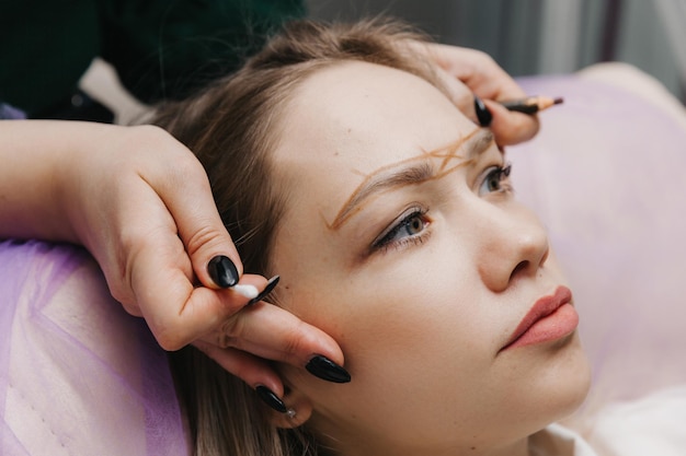 Girl with permanent makeup on her eyebrows The makeup artist makes markings with a brown pencil for eyebrow tattooing