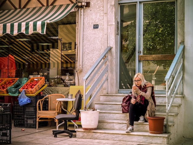 A girl with a phone is sitting on the steps of a shop selling vegetables and fruits in Greece