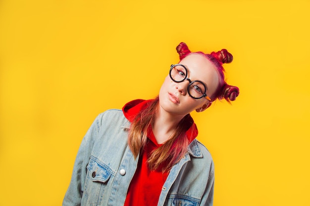 Girl with pink hair buns looking interested through round
glasses leaned teenager in red hoodie and jeans jacket isolated on
yellow background