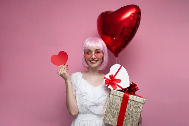 Girl with pink hair in a dress holds gifts and balloons and shows a valentine heart