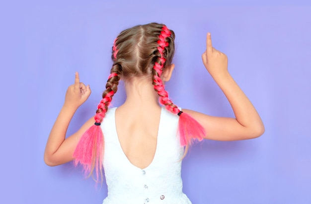 분홍색 땋은 머리가 카메라를 등지고 서 있는 소녀가 손으로 가리키고 보라색 스튜디오 배경에 공간을 복사 벽을 가리키는 어린 소녀의 후면 보기