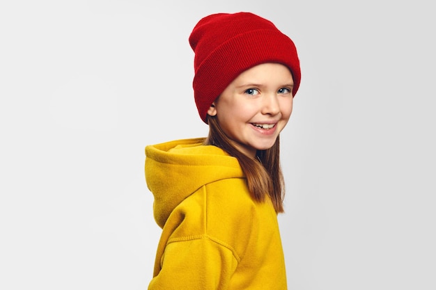 Girl with pleasant smile wears yellow hoodie and red hat stands against