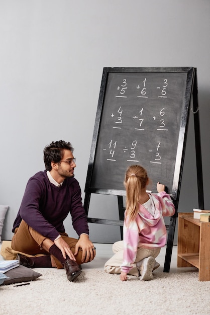 Girl with ponytail pointing at blackboard while learning to add and subtract numbers with tutor