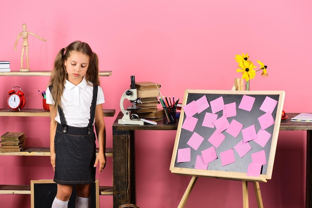 Girl with ponytails stands by blackboard with sticky notes desk with microscope and books Schoolgirl with sad face Back to school and childhood concept Kid and school supplies on pink background