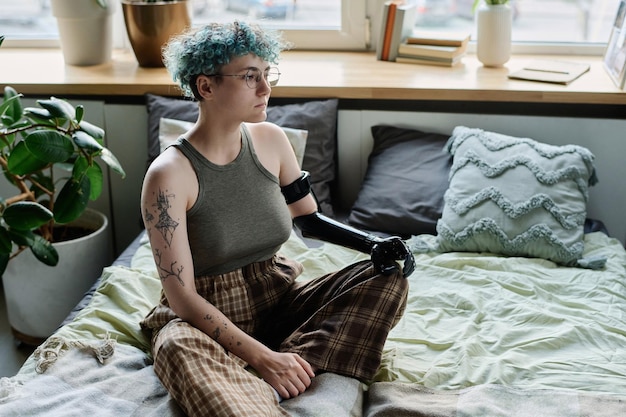 Girl with prosthetic arm sitting on bed with sad expression and thinking about something