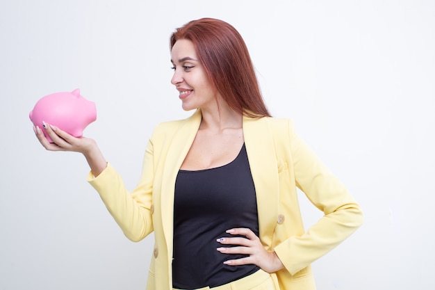 A girl with red hair holds a pink piggy bank in a yellow business suit