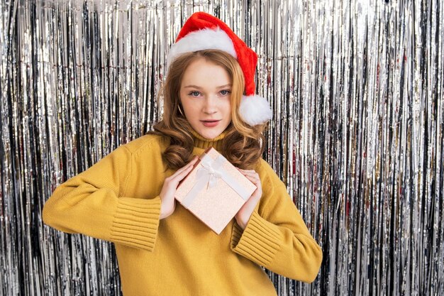 Girl with red hair and Santa hat holds a gift in her hand. She smiles and she is happy. Xmas